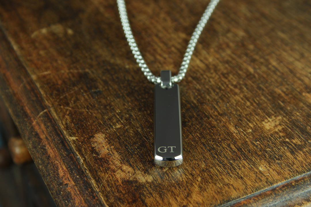 Black pendant with 'GT' initials on a chain, placed on a wooden surface.