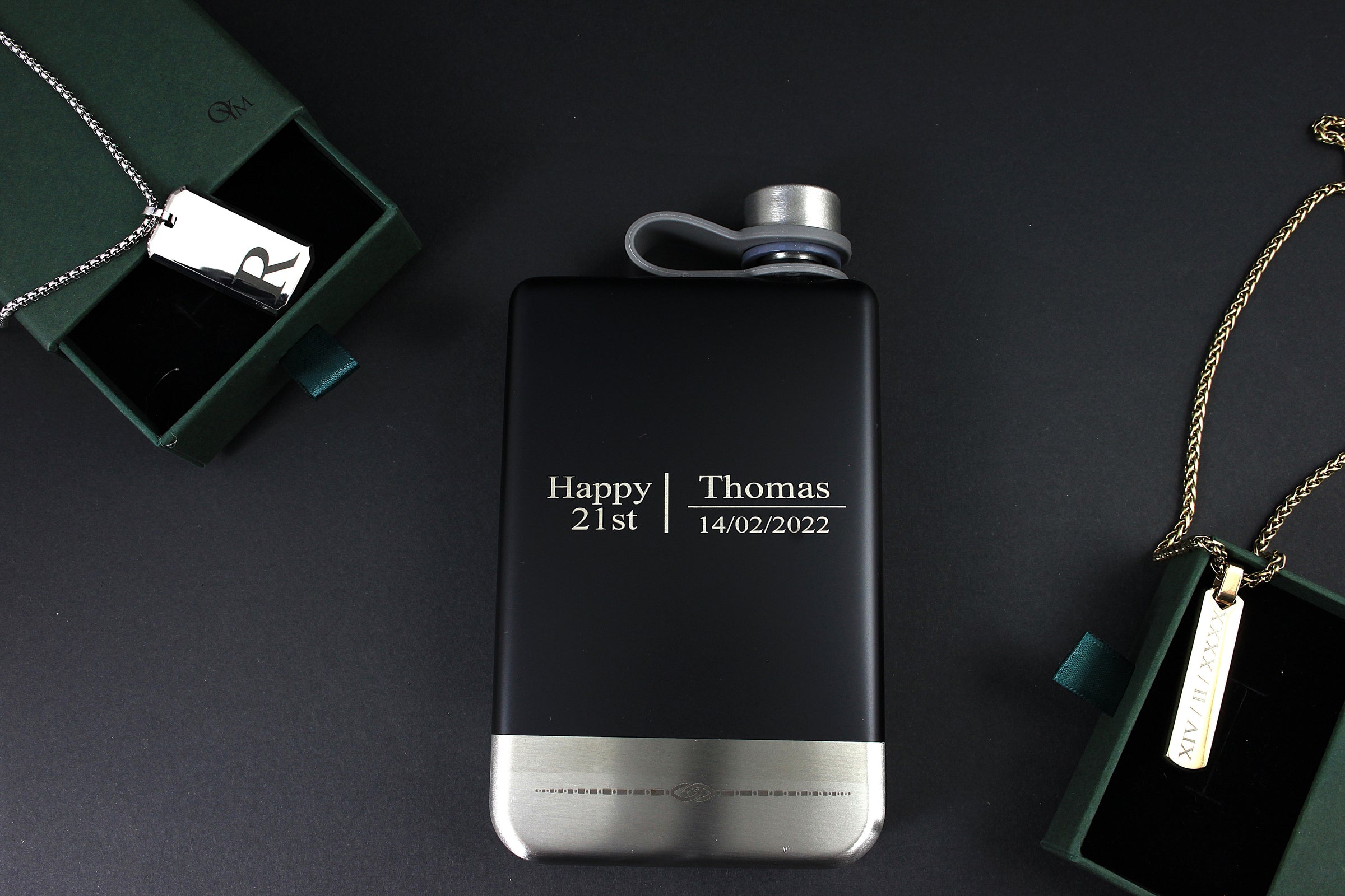 Black flask with engraved text on a dark surface, surrounded by green gift boxes and jewelry.