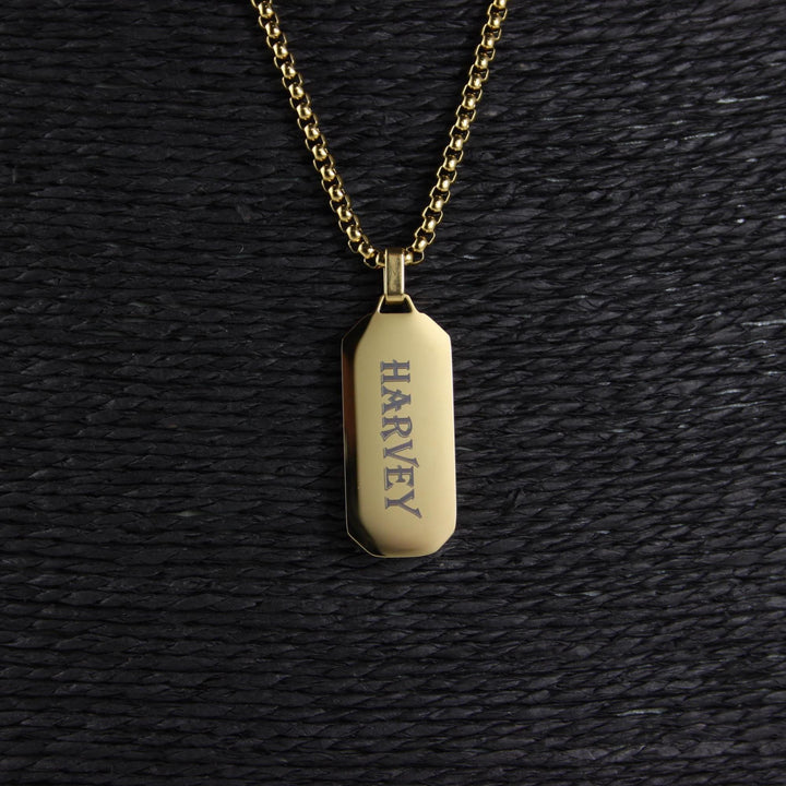Gold necklace with a tag engraved 'Harvey' on a black textured background