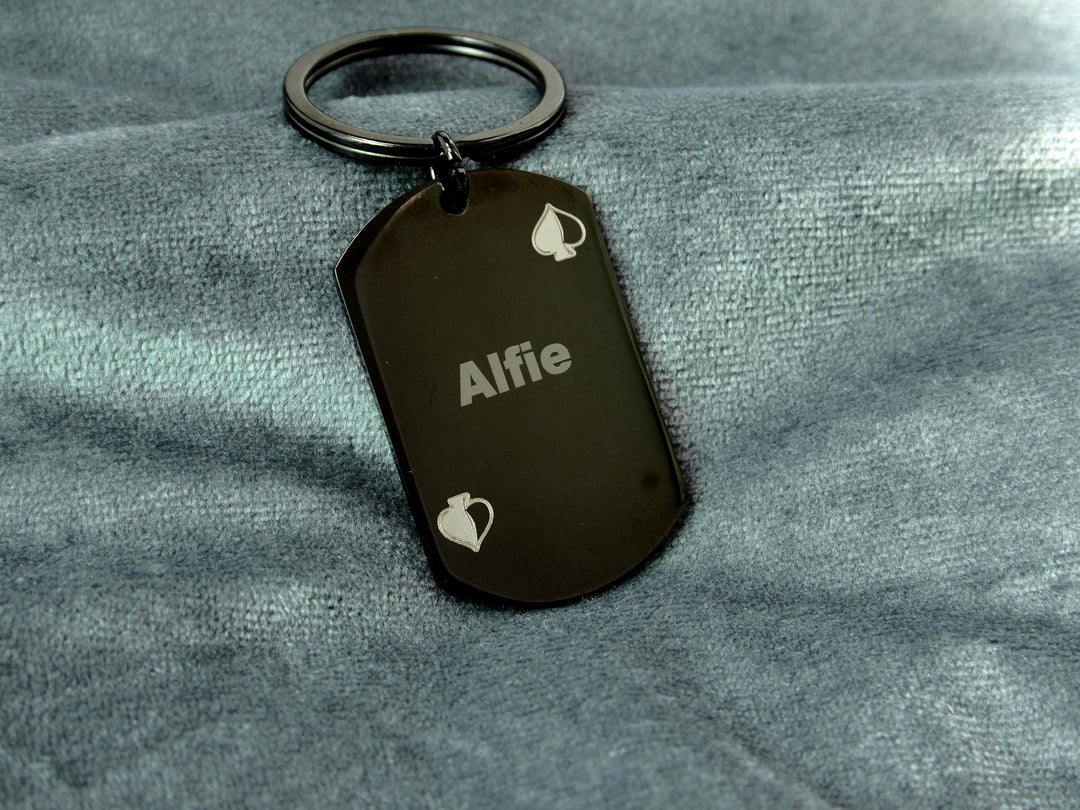 Black dog tag keychain with 'Alfie' engraved on a gray fabric background